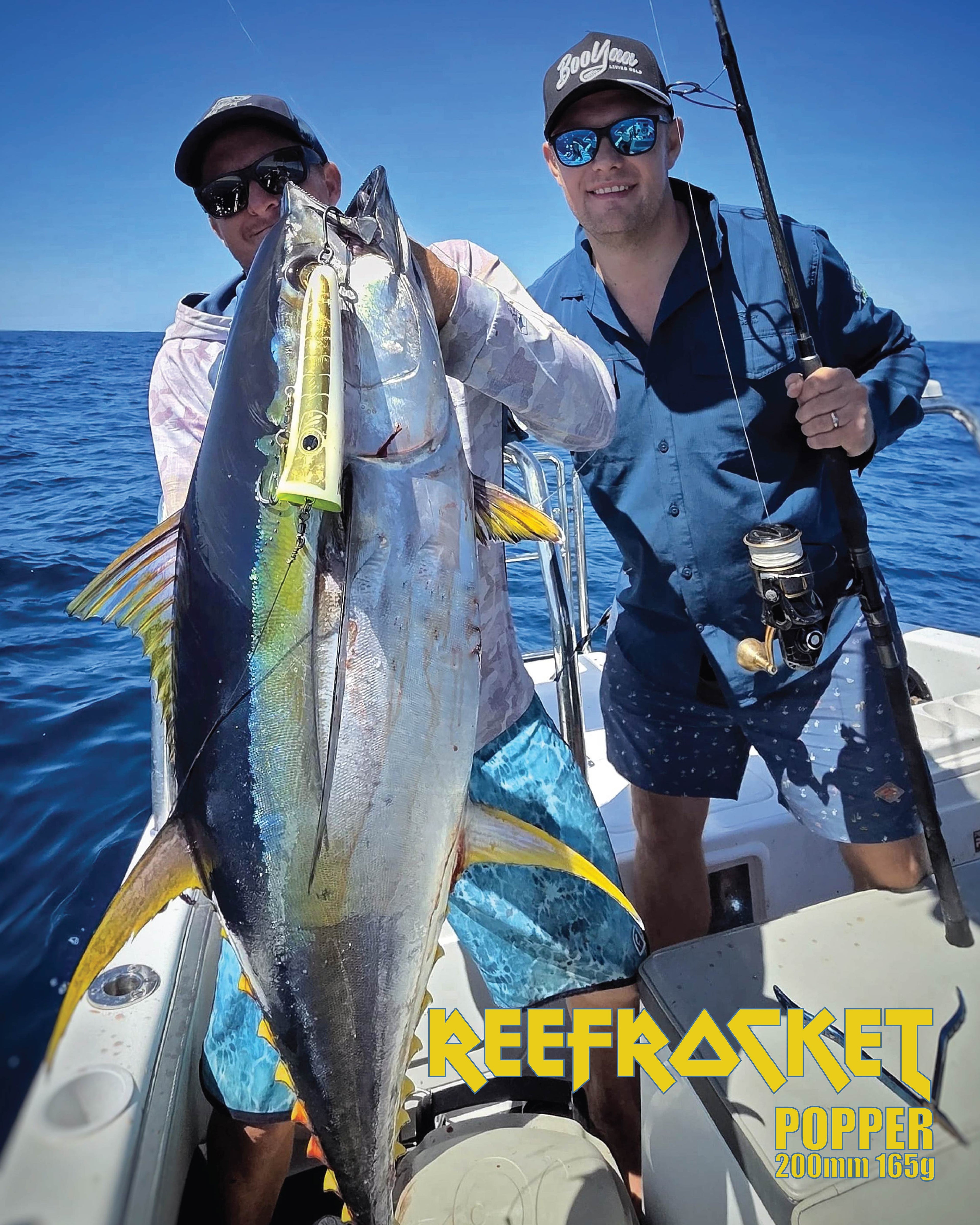 Two men on a boat holding a large yellow-fin tuna fish with a Reefsknot Popper lure attached, under a blue sky with ocean water in the background. The text 'REEFROCKET POPPER 200mm 4.